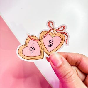 May include: A pink and gold heart-shaped locket with a pink ribbon and the letters "N" and "G" inside.