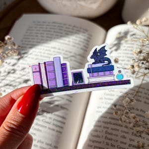Könnte beinhalten: Ein holografischer Aufkleber mit einem Miniatur-Bücherregal mit lila und weißen Büchern, einem kleinen gerahmten Bild und einer Drachenfigur. Eine kleine blaue Flaschen-Potion ist ebenfalls enthalten. Der Aufkleber wird über einem offenen Buch gehalten.