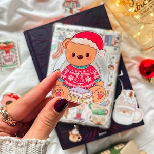 Könnte beinhalten: Ein klarer Aufkleber mit einem Cartoon-Bären, der einen roten Weihnachtspullover und einen Weihnachtsmannhut trägt und auf Büchern sitzt. Der Aufkleber wird vor einem Buch und anderen weihnachtlichen Aufklebern gehalten.