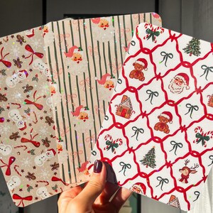 以下が含まれることがあります： 3つのクリスマスをテーマにしたカード。雪だるま、ジンジャーブレッドマン、リボンがベージュの背景に描かれたカード。ストライプの紙にサンタクロースの顔が描かれたカード。テディベア、リボン、クリスマスツリーが描かれたカード。