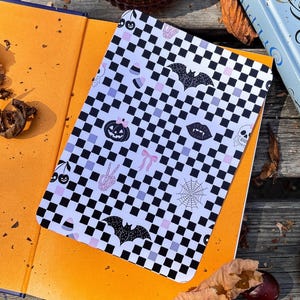 May include: A notebook with a black and white checkered pattern and Halloween-themed illustrations, including bats, pumpkins, and spiderwebs. The notebook is open on top of an orange book, with a candle and a book in the background.