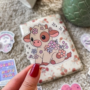 Puede incluir: Una pegatina con una vaca de dibujos animados con flores rosas y moradas. La vaca es blanca con manchas marrones y tiene la nariz rosa. La pegatina está sobre un fondo blanco.