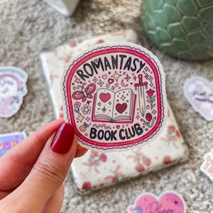 Könnte beinhalten: Ein rosa-weißer Aufkleber mit einem Buch, einem Schwert, einem Schmetterling und Herzen. Der Text auf dem Aufkleber lautet "Romantasy Book Club".
