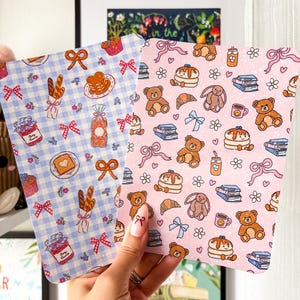 Può includere: Due carte decorative con motivi di cibo e orsetti. Una ha uno sfondo a quadretti blu con illustrazioni di pane, pancake e marmellata. L'altra è rosa con orsetti, pancake e libri. Entrambe le carte hanno fiocchi.