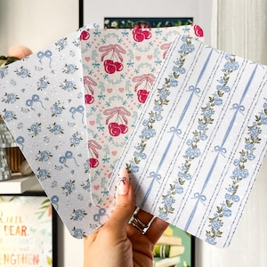 Può includere: Tre biglietti decorativi con motivi floreali e di ciliegie. I biglietti presentano disegni con rose blu, fiocchi e ciliegie su sfondo bianco. I biglietti sono tenuti in mano, mostrando la varietà dei motivi.