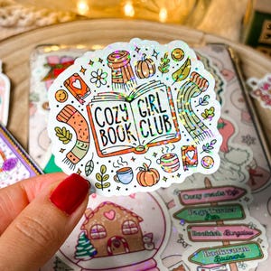 Könnte beinhalten: Ein weißer Aufkleber mit glitzernder Oberfläche, der ein offenes Buch mit dem Text "Cozy Girl Book Club" zeigt. Das Buch ist von Illustrationen von Herbstmotiven wie Kürbissen, Blättern und einer Tasse Kaffee umgeben.