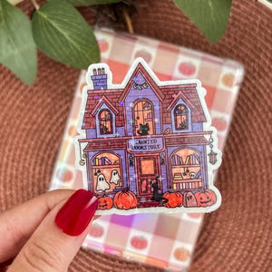 Peut inclure: Un autocollant représentant une librairie hantée violette et rouge avec des citrouilles, des fantômes et un chat noir. Le texte "Haunted Bookstore" est visible. L'autocollant est tenu par une personne avec du vernis à ongles rouge.