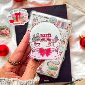 以下が含まれることがあります： ピンク色のベースとリボンが付いた、透明で丸いスノーグローブステッカー。内部には、お店、木、雪のあるお祭りシーンがあります。「My Holidays are Booked」と書かれたものなど、他のステッカーやオーナメントが背景に見えます。