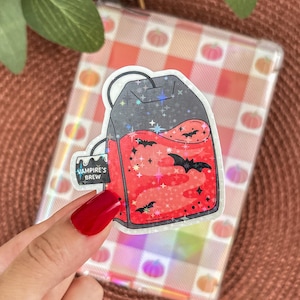 May include: A sticker depicting a tea bag filled with red liquid, decorated with black bats and stars. The tag reads "Vampire's Brew". The background is a clear, holographic surface with a pumpkin pattern.