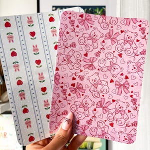 Könnte beinhalten: Zwei dekorative Karten mit gemusterten Designs. Eine Karte hat einen hellblauen Hintergrund mit vertikalen Streifen, Hasen, Herzen und Kirschen. Die andere Karte hat einen rosa Hintergrund mit Teddybären, Schleifen, Rosen und Herzen.