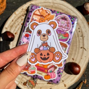 Può includere: Un adesivo a tema Halloween con un orso panda vestito da fantasma, che tiene in mano una zucca piena di caramelle. L'adesivo è su uno sfondo viola con altri adesivi e una superficie di legno.