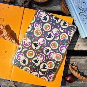 May include: A clear case with a Halloween-themed pattern featuring black cats, ghosts, and jack-o'-lanterns on a dark background. The case is resting on an open orange book. Candy corn accents the design. A candle and a book are in the background.