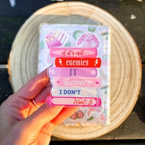 May include: A stack of pink and red book-shaped stickers with text that reads "if it's not enemies", "II", "Lovers", "I DON'T", and "Want It". The stickers are held in front of a clear background with other stickers.