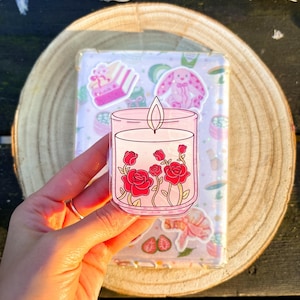 Könnte beinhalten: Ein klarer Aufkleber mit einer rosa Kerze mit roten Rosen. Der Aufkleber wird vor einer transparenten Hülle gehalten, die mit pastellfarbenen Aufklebern verziert ist, darunter ein rosa Hase und ein Kuchen. Der Hintergrund umfasst eine Holzscheibe und Schnee.
