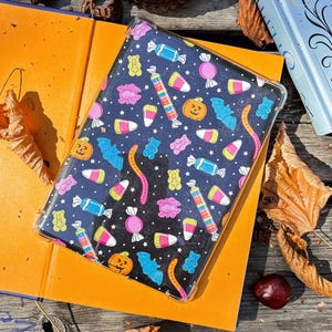 May include: A black tablet case with a colorful Halloween candy pattern. The design includes pumpkins, bats, candy corn, gummy bears, and wrapped candies. The case is on top of an open orange book.