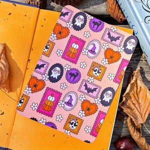 May include: A pink notebook with a Halloween-themed pattern featuring pumpkins, bats, ghosts, and spiders. The notebook is open on top of a book with orange pages. A candle and a book are in the background.