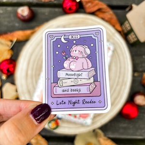 Peut inclure: Autocollant rectangulaire avec un ours en peluche de dessin animé portant un casque, assis sur des livres. Le sticker a une bordure violette avec le texte "XII", "Moonlight and books", et "Late Night Reader". L'arrière-plan est violet clair avec des étoiles et un croissant de lune.