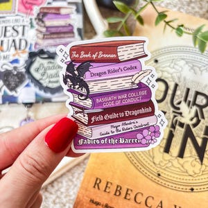 May include: A sticker featuring a stack of fantasy books with titles like "The Book of Brennan" and "Dragon Rider's Codex." A small black dragon is perched on the books. The sticker has a white border and is held by a hand with red nail polish.