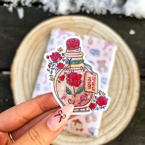 May include: A sticker featuring a potion bottle with a red rose inside. The bottle has a red stopper and a tag that reads "Romance Potion." Red roses and hearts decorate the bottle. The sticker is on a wooden surface.