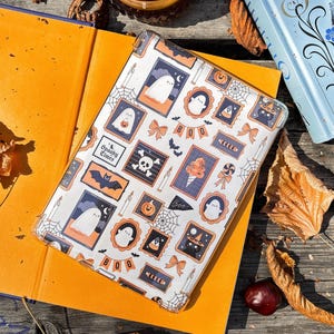 May include: A clear phone case with a Halloween-themed design featuring ghosts, pumpkins, bats, and the word "Boo!" The case is on top of an open orange book. A lit candle, a blue book, and fall leaves are also in the image.