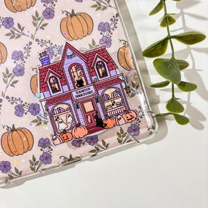 Puede incluir: Funda transparente para teléfono con diseño de Halloween. La funda presenta un patrón repetido de calabazas naranjas y flores moradas. Una ilustración central representa una librería encantada morada con calabazas y fantasmas.