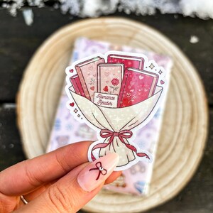 Può includere: Un adesivo raffigurante un bouquet di libri con copertine rosa e rosse, avvolto in carta bianca e legato con un fiocco rosso. Il testo "Romance Reader" è visibile su una piccola carta. Cuori e stelle decorano il design.