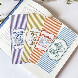 Peut inclure: Quatre marque-pages colorés avec des rayures verticales et des illustrations fantaisistes. Les marque-pages présentent des phrases comme "Book Girl Summer" et "Silly Goose on the Loos". Chaque marque-page a un design et une palette de couleurs uniques.