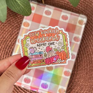 Może przedstawiać: Kolorowa naklejka z napisem "cozy bookish weekend ADMIT ONE" przedstawiająca stos książek, kubek, czapkę i dynie. Naklejka jest trzymana przed przezroczystym, opalizującym etui z wzorem dyni.