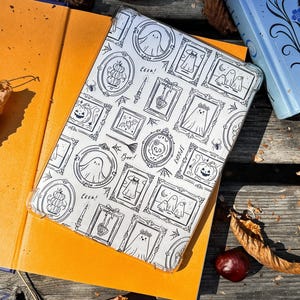 Puede incluir: Una funda transparente para teléfono con un patrón en blanco y negro con temática de Halloween que presenta fantasmas, calabazas y la palabra "Eek!". La funda está sobre un libro naranja abierto, con un libro azul y follaje de otoño en el fondo.