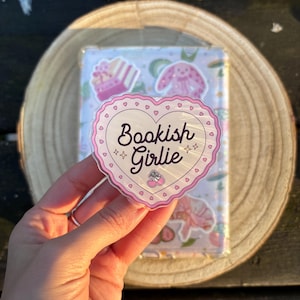 Könnte beinhalten: Ein herzförmiger Aufkleber mit dem Schriftzug "Bookish Girlie" in Schreibschrift, umgeben von einem rosa Rand mit Herzen. Der Aufkleber wird über einem Notizbuch mit rosa und weißen Illustrationen gehalten.