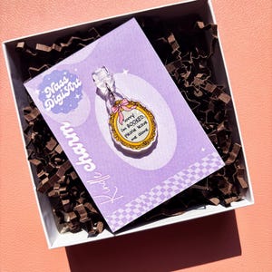 Peut inclure: Un charm transparent avec un cadre doré et le texte "sorry I'm BOOKED please leave me alone" sur une carte violette. Le charm est dans une boîte blanche avec du papier de remplissage marron, sur fond pêche. Le charm est de Nass Digi Art.