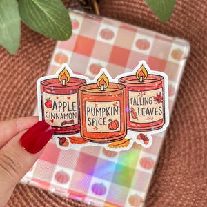 Peut inclure: Trois autocollants de bougies sur le thème de l'automne, avec les parfums Pomme Cannelle, Épices de Citrouille et Feuilles Mortes. Chaque autocollant présente une illustration de bougie avec une flamme et des éléments décoratifs. Les autocollants sont sur un fond transparent.