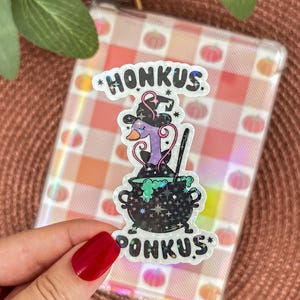 Könnte beinhalten: Ein klarer Aufkleber mit dem Text "HONKUS PONKUS" und einer Cartoon-Ente mit Hexenhut, die einen brodelnden Kessel umrührt. Der Kessel hat ein holografisches Design und grüne Flüssigkeit. Der Hintergrund ist eine transparente Handyhülle mit einem rosa-weißen Vichy-Muster.