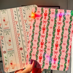 以下が含まれることがあります： クリスマスをテーマにした2冊のノート。1冊は、ガチョウ、リボン、そして「MERRY CHRISTMAS」の文字が入ったクリーム色の背景です。もう1冊は、くるみ割り人形、リボン、ガーランドが入ったピンク色の背景です。どちらも縦模様です。