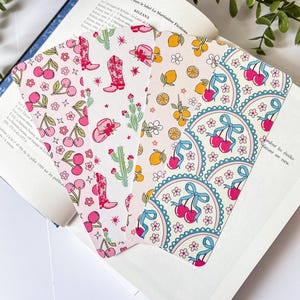 Peut inclure: Quatre marque-pages décoratifs avec des motifs colorés sont présentés dans un livre ouvert. Les motifs comprennent des cerises, des bottes de cowboy, des citrons et un motif floral. Les marque-pages sont sur fond blanc.