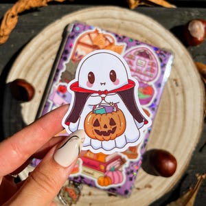 May include: A Halloween sticker of a friendly ghost wearing a cape and holding a pumpkin. The sticker is on a purple background with other Halloween illustrations. The sticker is held by a person. The background includes conkers and leaves.