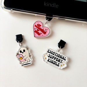 Peut inclure: Trois breloques différentes pour une liseuse Kindle. La première breloque est un cœur rose avec le texte "Bookish Soul" en rouge. La deuxième breloque est un fantôme tenant un livre avec le texte "Spooky AF" sur la couverture. La troisième breloque est un nuage blanc avec le texte "Emotional Support Kindle" en noir.