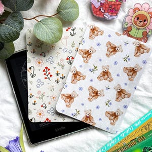 May include: Kindle e-reader with two decorative covers. One cover has a floral pattern with red and white flowers. The other cover features teddy bears and blue flowers. Two decorative pins and a stack of books are also visible.