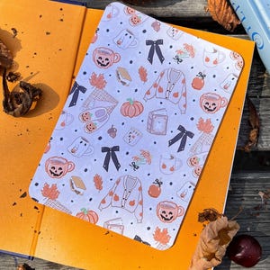 Pode incluir: Um bloco de notas retangular branco com um padrão repetido de abóboras, canecas, cardigans e laços. O bloco de notas está em cima de um livro laranja, com um livro azul e folhas de outono no fundo. O tema é o outono.