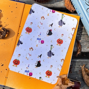 Può includere: Un taccuino a tema Halloween con un motivo di oche in costume, tra cui streghe, zucche e cowboy. Il taccuino è aperto, rivelando un interno arancione. Un libro e una candela sono visibili sullo sfondo.