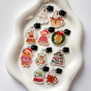 Peut inclure: Une collection de breloques sur le thème de Noël avec divers motifs, dont le Père Noël, des rennes, des boules à neige, un ours en peluche, un chat dans une couronne et un bonhomme en pain d'épices. Chaque breloque a un fermoir noir. Les breloques sont présentées sur un plat blanc aux bords ondulés.