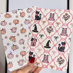 Può includere: Due biglietti decorativi a tema Halloween. Uno presenta zucche e fiori, l'altro gatti neri, cappelli da strega e fiocchi. Entrambi i biglietti hanno uno sfondo bianco con accenti rosa e glitter.