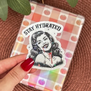 Può includere: Un adesivo con un'illustrazione retrò di una donna con zanne e sangue che gocciola dalla bocca, con la scritta "Stay Hydrated". L'adesivo ha un effetto olografico ed è ambientato su uno sfondo a quadri con motivi di zucca.
