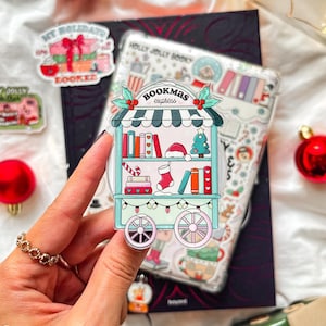 Könnte beinhalten: Ein farbenfroher Aufkleber mit einem weihnachtlichen Bücherwagen mit dem Text "Bookmas Express". Der Wagen ist mit Büchern, einem Weihnachtsmannhut, einem Weihnachtsbaum und Lichterketten geschmückt. Weitere Aufkleber und Ornamente sind im Hintergrund zu sehen.
