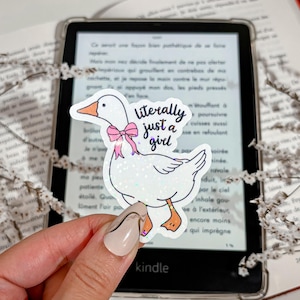 Puede incluir: Una pegatina blanca con un ganso de dibujos animados con un lazo rosa y el texto "literally just a girl". La pegatina se sostiene frente a un lector Kindle, con un libro en el fondo.