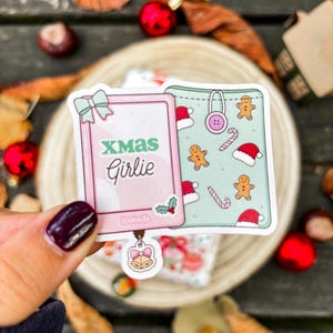 Op de afbeelding: Twee stickers met kerstthema. Eén is roze met de tekst "XMAS Girlie" en een strik. De andere is mintgroen met peperkoekmannetjes, zuurstokken en kerstmutsen. Een kleine belletjesbedel hangt aan de roze sticker.