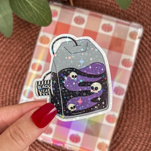 Könnte beinhalten: Ein holografischer Aufkleber in Form eines schwarzen Teebeutels mit dem Schriftzug "BLACK TEA" auf dem Etikett. Das Teebeutel-Design zeigt lila Wirbel, Sterne und drei kleine Totenköpfe. Der Aufkleber wird von einer Hand mit rotem Nagellack gehalten.
