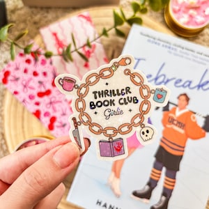 Op de afbeelding: Een sticker met een gouden kettingontwerp en de woorden "Thriller Book Club Girlie". De sticker bevat kleine illustraties van een koffiekopje, een hart, een mes, een boek en een schedel. Een boek en een kaars staan op de achtergrond.