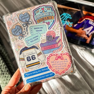 Peut inclure: Coque de téléphone transparente avec un motif floral et divers autocollants. Les autocollants comprennent des motifs sur le thème du hockey, un maillot avec le numéro 66 et du texte qui dit "In My Hockey Romance" et "You're killing me, Wellsey."