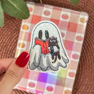 Peut inclure: Autocollant holographique représentant un fantôme lisant un livre avec un chat noir. Le fantôme est blanc avec des yeux noirs et tient un livre rouge. Le chat est noir et tient une pile de livres. Le fond est un motif à carreaux rose et orange.